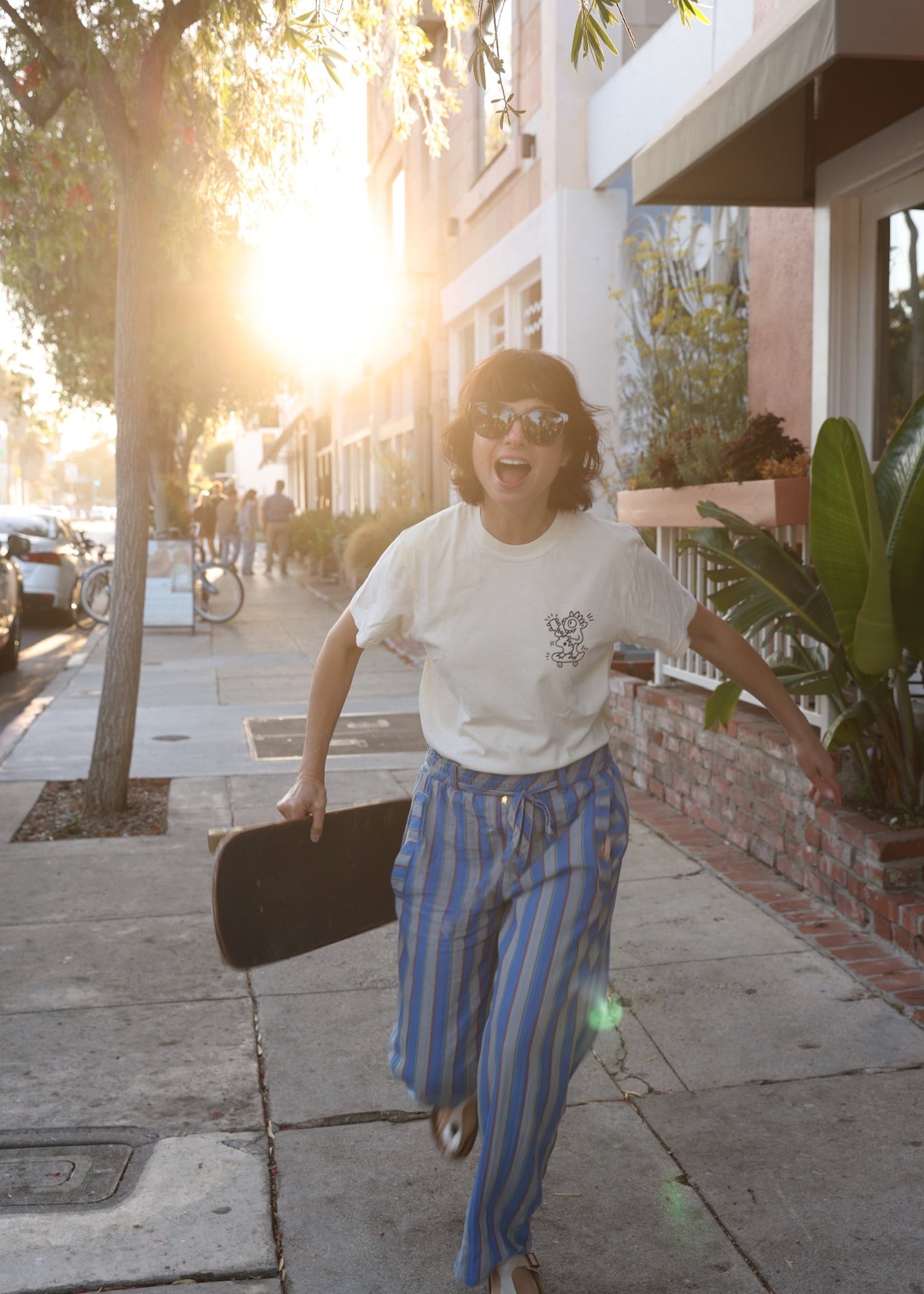 Person walking on a sidewalk with a skateboard, wearing a white t-shirt and blue striped pants.