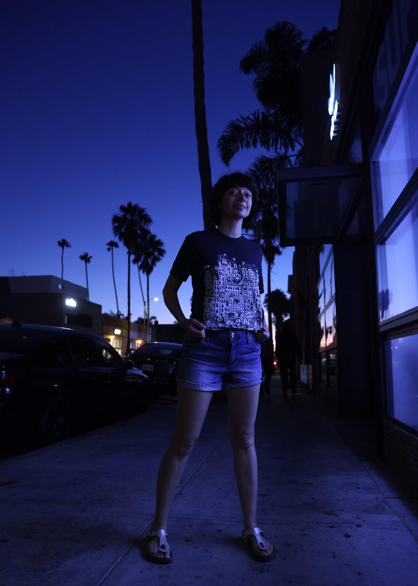 Person standing on a street at night with palm trees and buildings in the background