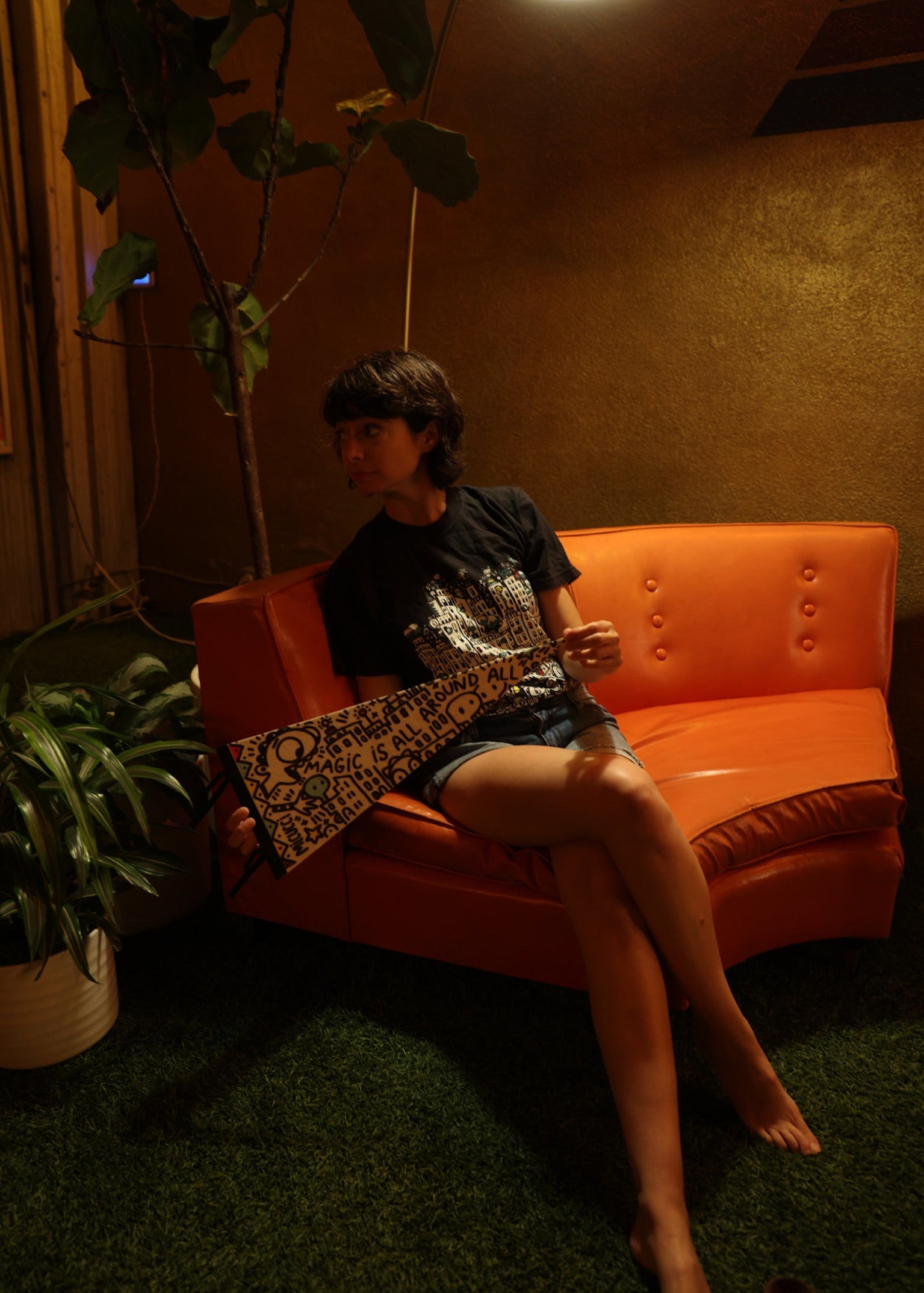 Person sitting on an orange couch in a dimly lit room with plants.